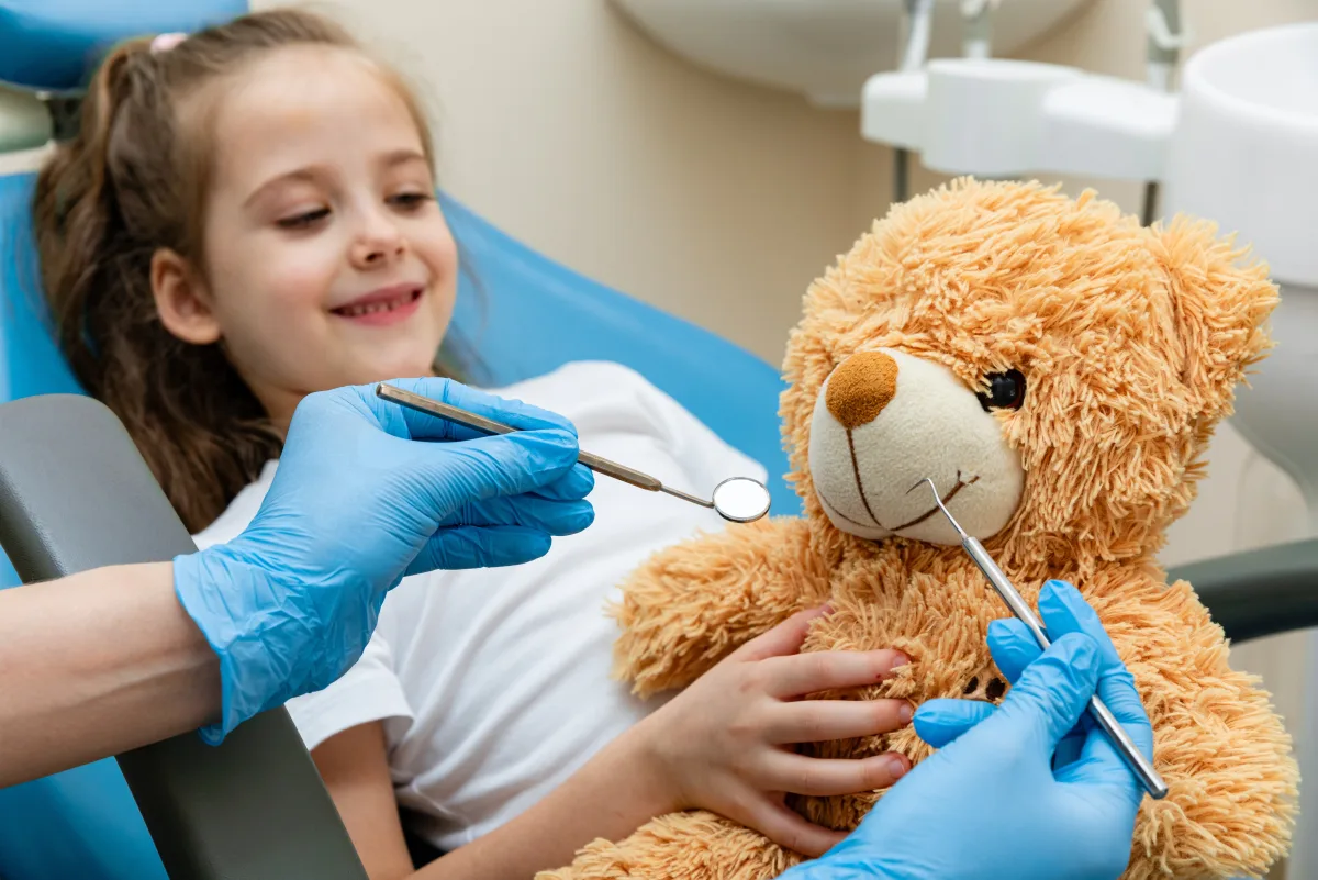 Child in a dental chair smiling while the dental team demonstrates a mirror and explorer on a teddy bear during a tell-show-do first visit